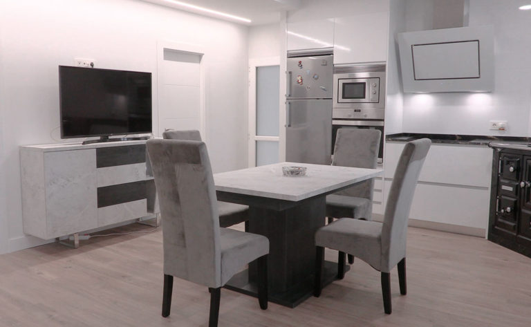 Vista frontal de cocina blanca en ele con mesa de comedor en la zona central, mueble para TV y electrodomésticos integrados