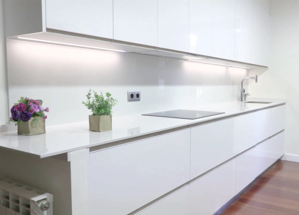 Vista lateral de cocina blanca con encimera de porcelánico blanco con vitrocerámica y fregadero con grifo moderno, enchufes y muebles altos con iluminación led en los bajos