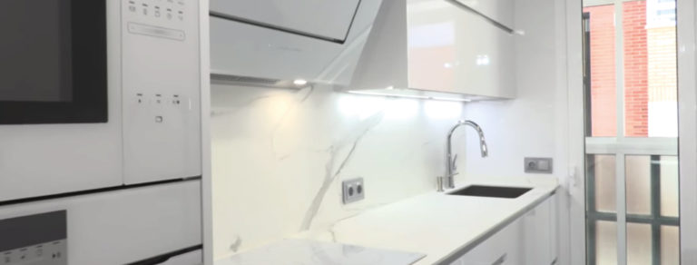 Vista lateral de cocina en color blanco con paredes en material similar al mármol, fregadero y electrodomésticos integrados y campana extractora y placa blancas