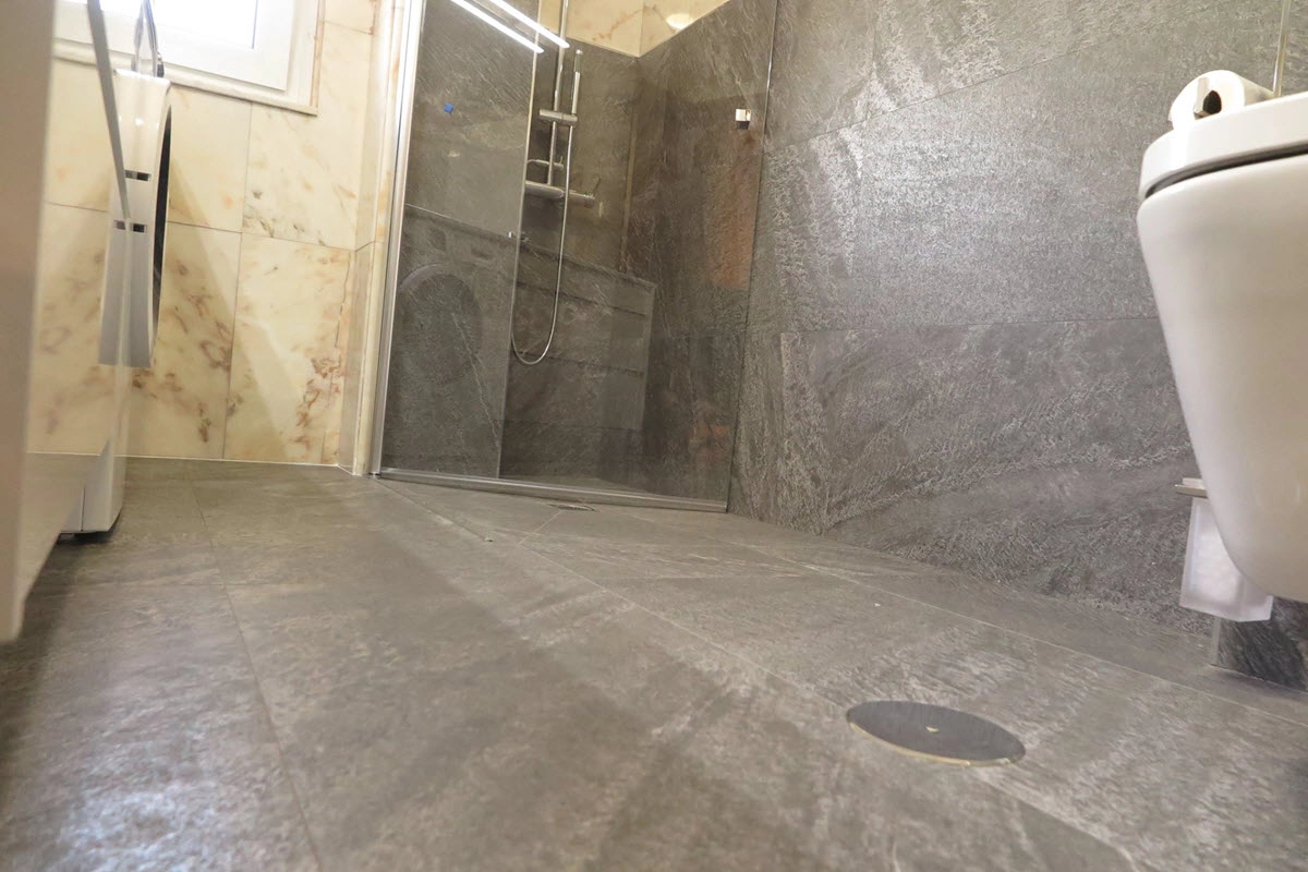 Vista desde el suelo de baño con azulejos de suelo y parte de la pared en color gris y otra en tonos anaranjados con inodoro, lavadora, mueble de lavabo y ducha