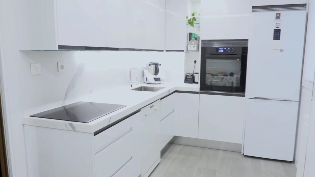 Cocina blanca pequeña en forma de ele con luz en los bajos de los muebles altos, nevera y horno integrados, placa de inducción negra y fregadero