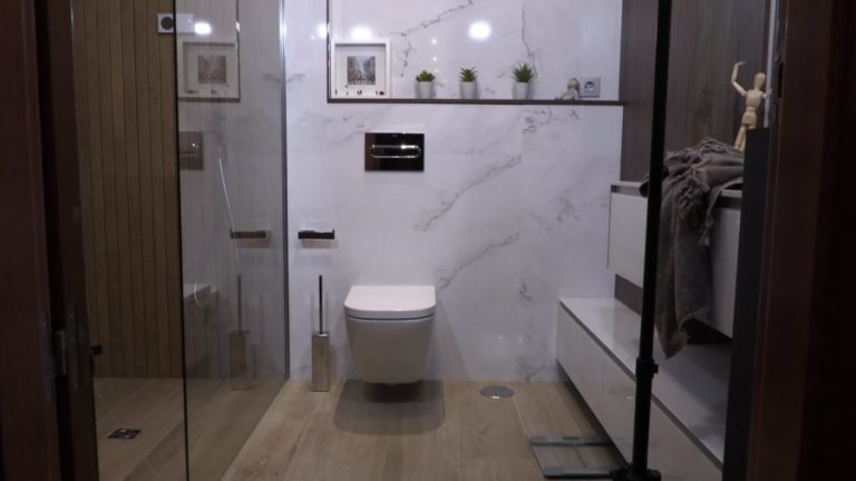 Vista frontal de baño oscuro en tonos blancos y detalles en madera con inodoro suspendido blanco, mueble de baño blanco y ducha de madera
