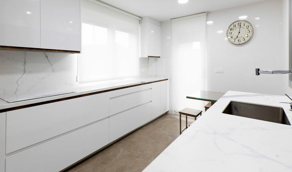 Vista lateral de cocina blanca moderna con detalles en madera con dos frentes con encimeras blancas marmoladas y espacio de almacenamiento
