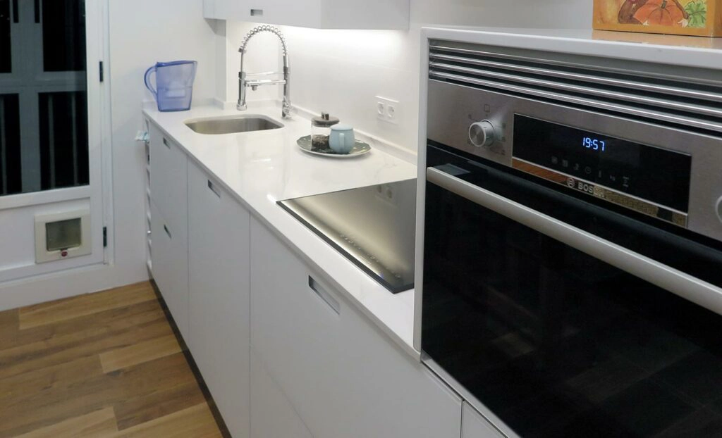 Vista lateral de cocina blanca pequeña con puertas de mobiliario de aluminio en cristal, fregadero, vitrocerámica y horno integrado