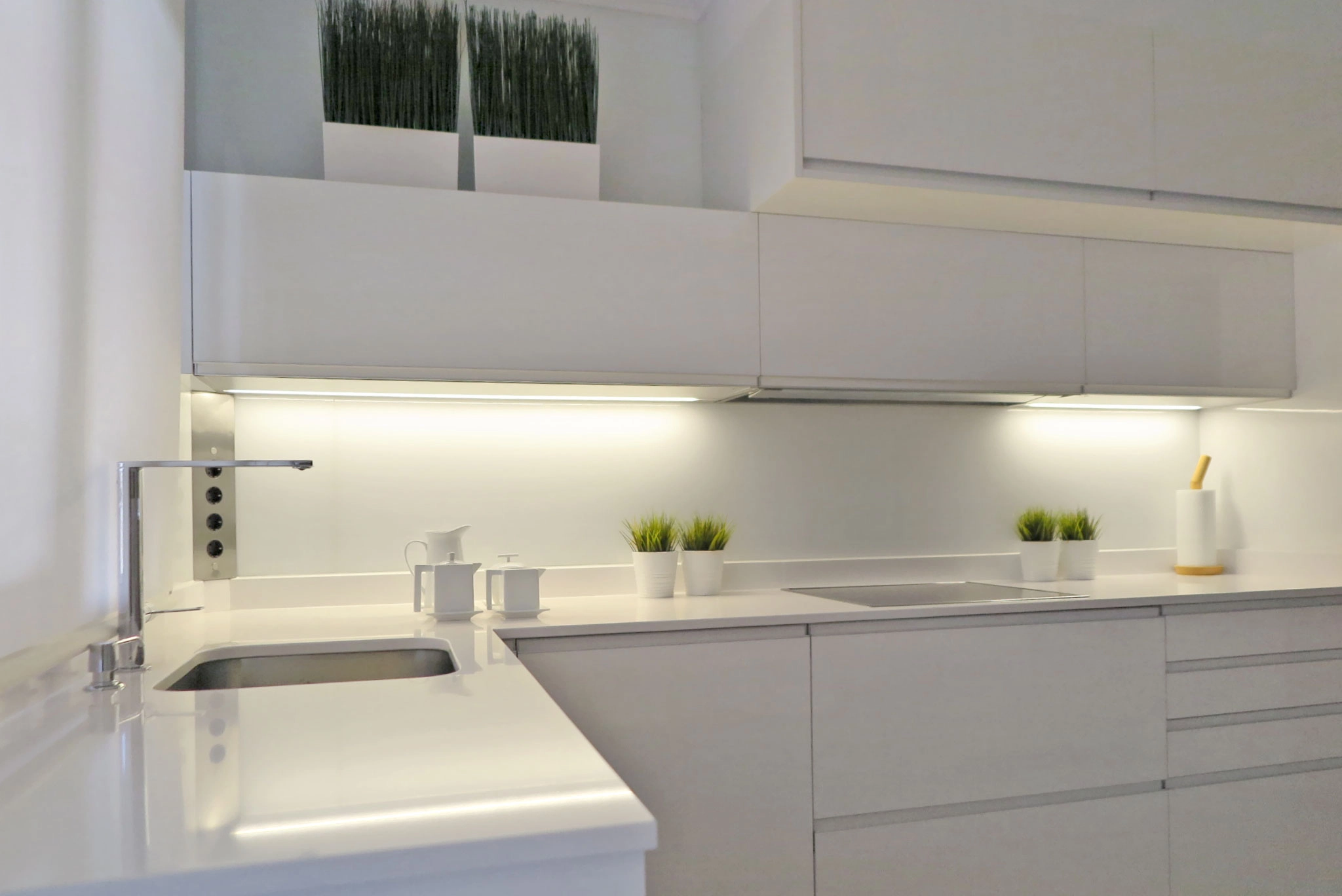Detalle de cocina blanca en ele con fregadero, vitrocerámica, iluminación led en la base de los muebles altos y tirador uñero integrado en todo el mobiliario
