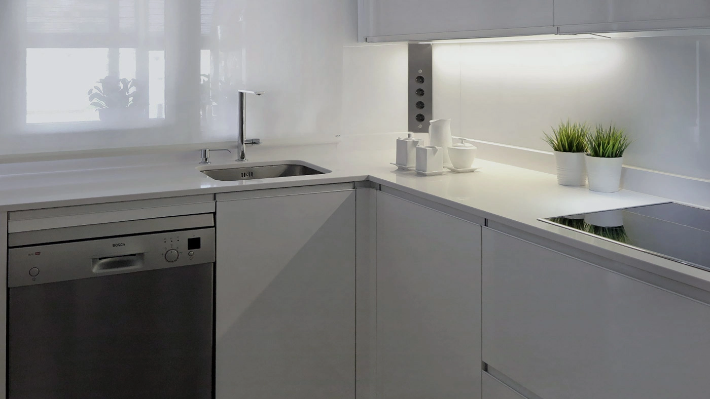 Vista frontal de cocina blanca con encimera en ele blanca con fregadero y vitrocerámica, lavavajillas y muebles bajos con tirador uñero integrado