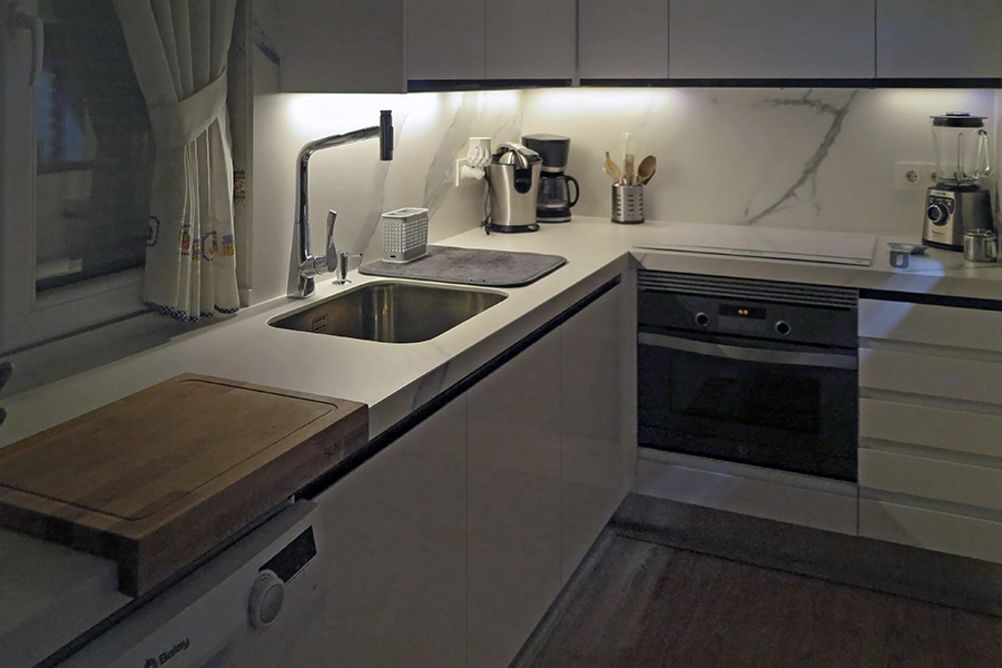Vista noctura de cocina moderna en color blanco con distribución en ele. Vista de zona de trabajo, fregadero y horno bajo encimera