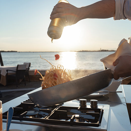 Cocinero preparando pasta al aire libre en cocina modular en una terraza con el mar al fondo