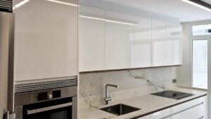 Frente de cocina moderna blanca con muebles altos, bajos y columna con horno, en color blanco brillo y tirador uñero en melamina roble dorado