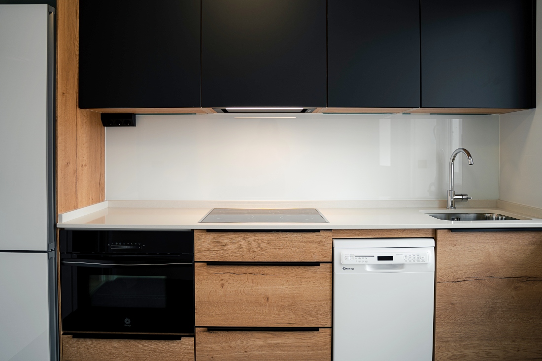 Vista frontal de cocina combinada en negro y madera con encimera blanca con fregadero y vitrocerámica y electrodomésticos integrados