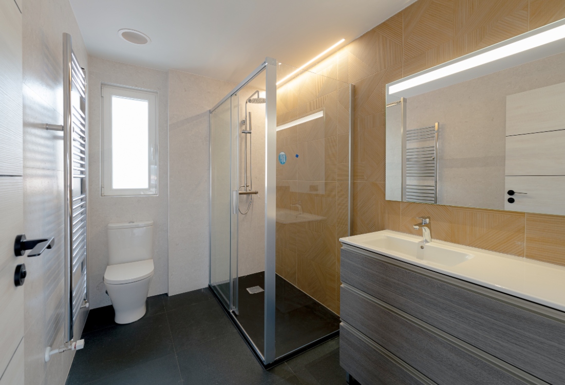 Vista lateral de cuarto de baño en tonos cálidos con inodoro, toallero, ducha con mampara transparente y mueble de baño amplio con lavabo y espejo rectangular sobre pared