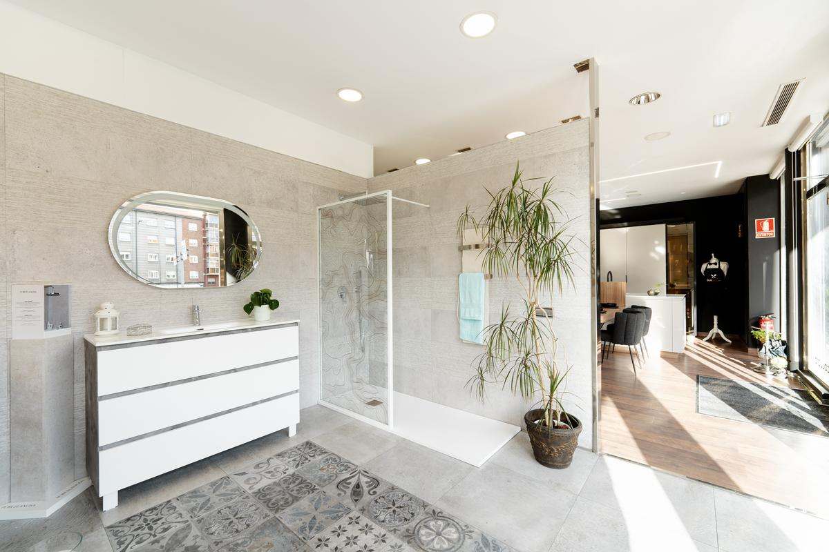 Exposición de baño moderno en tonos grises con mueble blanco y gris con lavabo incorporado y espejo ovalado encima y ducha a la derecha con mampara fija