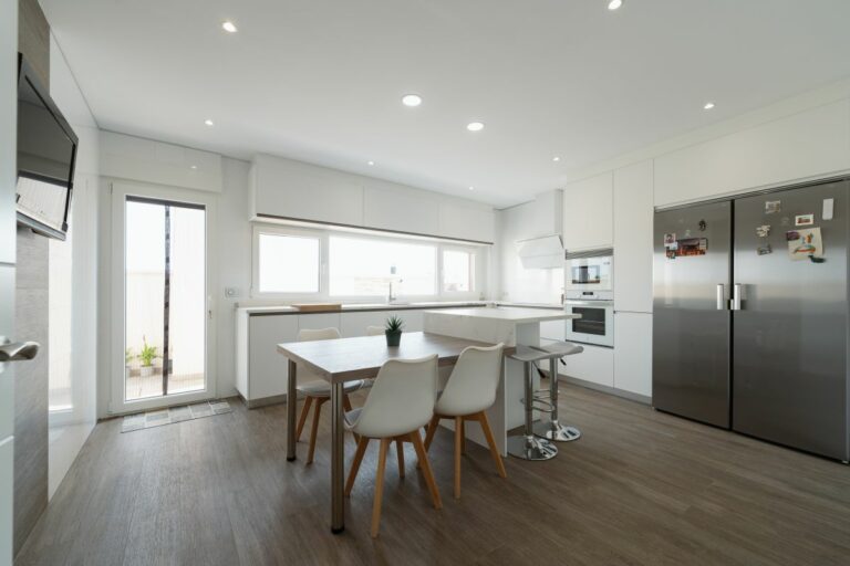 Vista general de una cocina moderna con mobiliario blanco sin tiradores, isla central con barra, mesa integrada con sillas blancas, doble frigorífico en acero inoxidable y gran entrada de luz natural