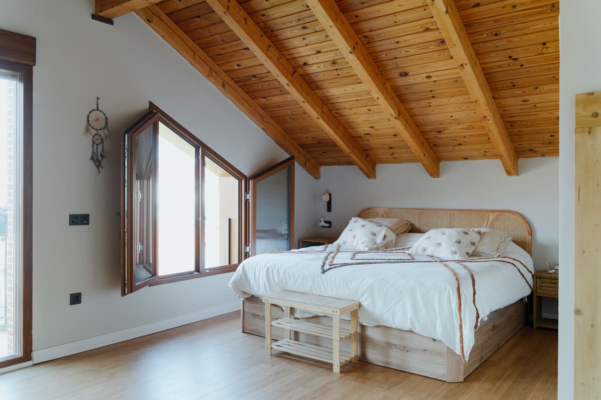 Dormitorio bajo cubierta con techo de madera vista, cama y ventanas que aportan luz natural.