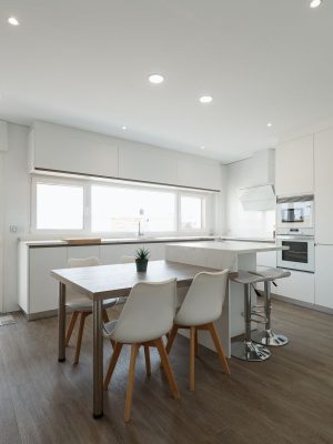 Vista general de una cocina moderna con mobiliario blanco sin tiradores, isla central con barra, mesa integrada con sillas blancas, doble frigorífico en acero inoxidable y gran entrada de luz natural